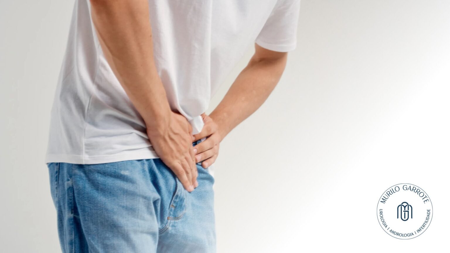 Homem vestindo camiseta branca e jeans, segurando a região da virilha, sugerindo desconforto ou preocupação.